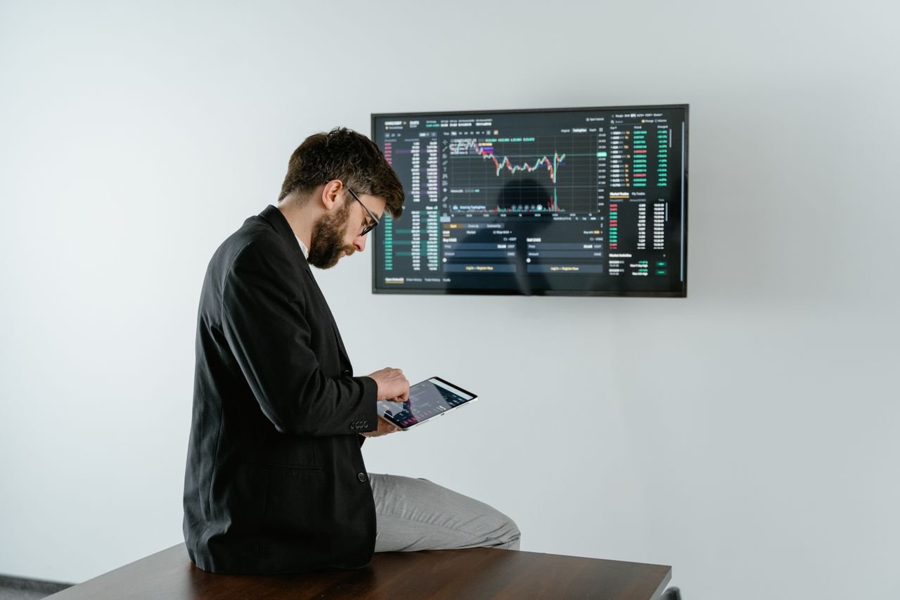 Young man in office analyzing financial graphs on a tablet with stock market screen displayed.
